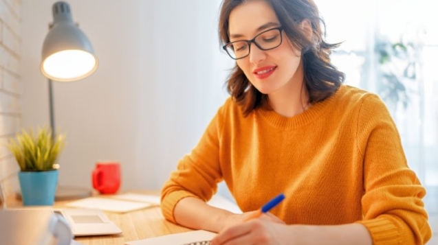 Smiling woman in glasses writing notes at a desk by a window