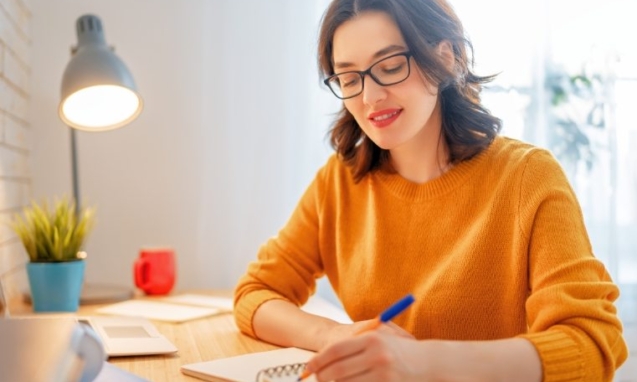 Smiling woman in glasses writing notes at a desk by a window