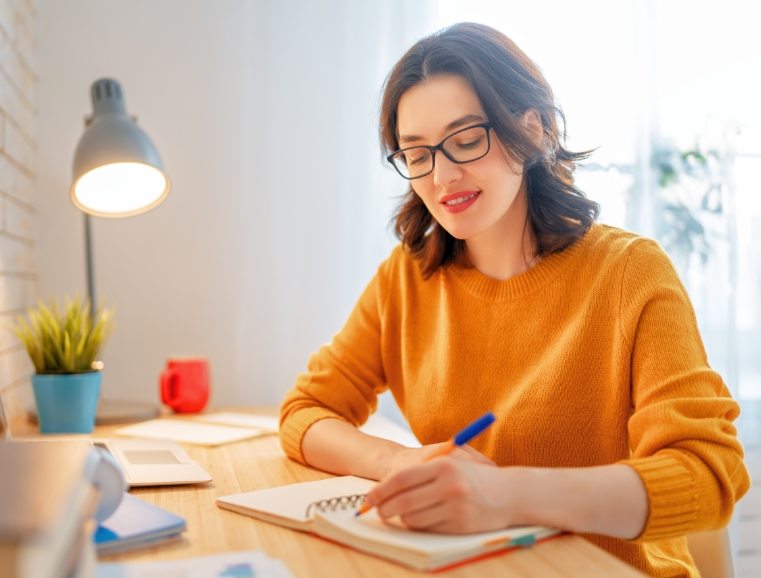 Smiling woman in glasses writing notes at a desk by a window